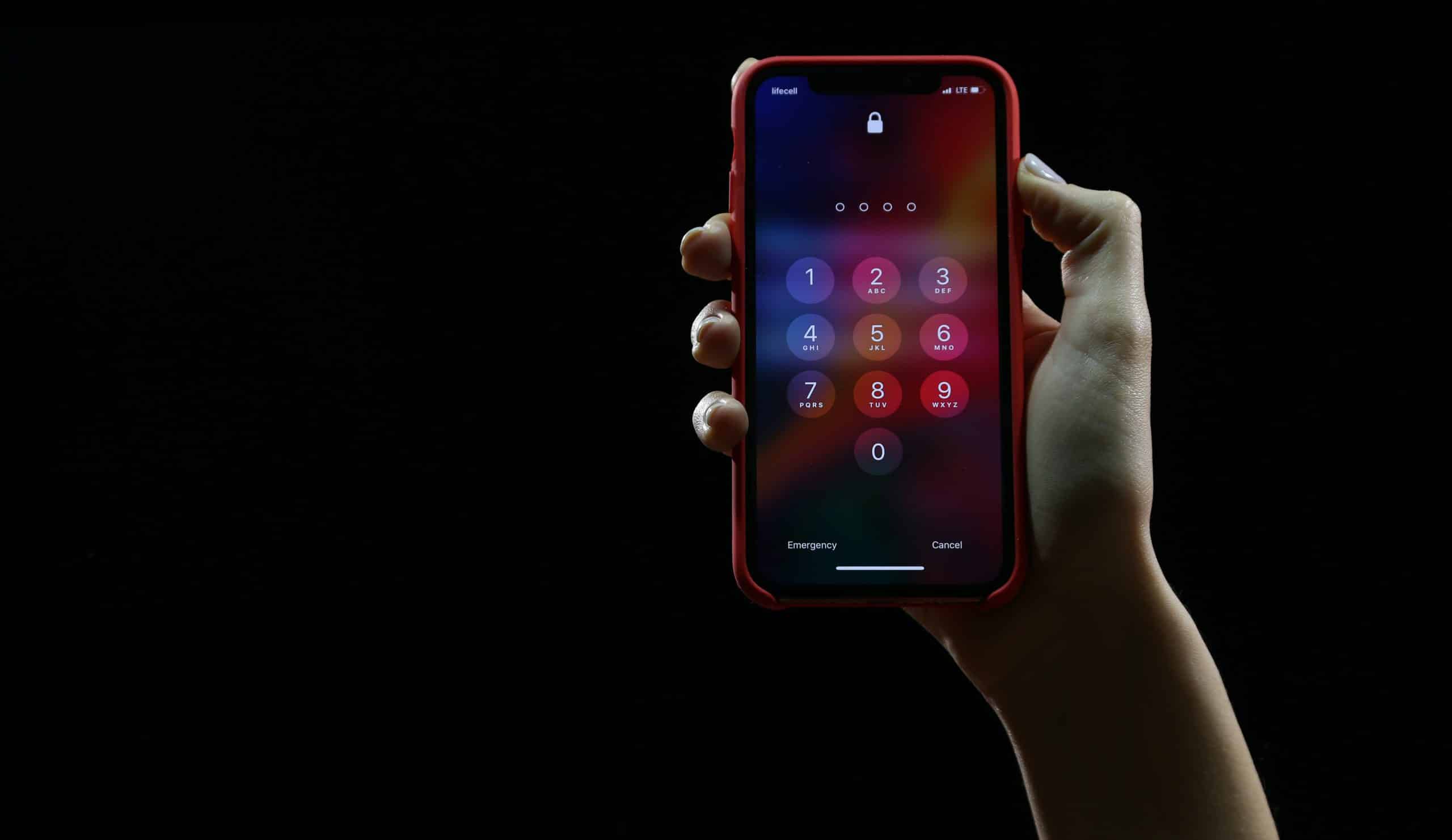 A hand holds a smartphone with a red case against a black background. The phone's screen shows a lock screen with a numeric keypad for entering a passcode, highlighting the importance of password management. The numbers 1 to 9 and 0 are visible, along with options for Emergency and Cancel at the bottom.