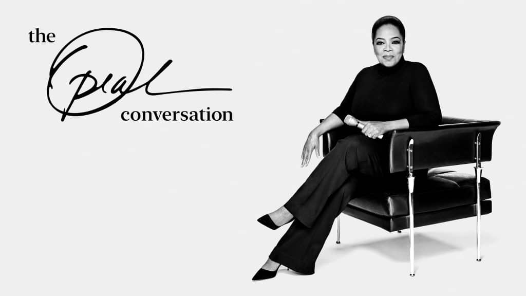 A person wearing a black outfit sits on a modern black chair, crossing their legs, and looking towards the camera with a calm expression. The text beside them reads, "The Oprah Conversation" with a stylized signature. This new talk show featuring Oprah Winfrey is available on Apple TV+. The background is plain white.