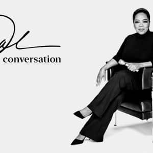 A person wearing a black outfit sits on a modern black chair, crossing their legs, and looking towards the camera with a calm expression. The text beside them reads, "The Oprah Conversation" with a stylized signature. This new talk show featuring Oprah Winfrey is available on Apple TV+. The background is plain white.