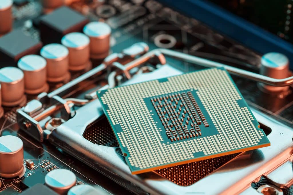 Close-up view of a next-generation Intel processor chip partially inserted into a motherboard socket. The intricate pins on the underside of the chip are clearly visible, while surrounding capacitors and circuit pathways are slightly out of focus in the background.