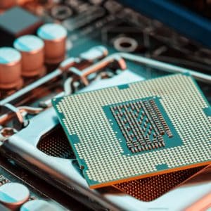 Close-up view of a next-generation Intel processor chip partially inserted into a motherboard socket. The intricate pins on the underside of the chip are clearly visible, while surrounding capacitors and circuit pathways are slightly out of focus in the background.