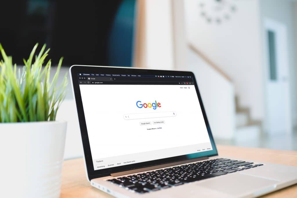 A laptop displaying the Google homepage is on a wooden desk. Next to it on the left is a potted plant with green leaves. The background shows a modern, bright home interior with white walls and a staircase, all highlighting the sleek design often seen in macOS devices.