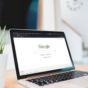 A laptop displaying the Google homepage is on a wooden desk. Next to it on the left is a potted plant with green leaves. The background shows a modern, bright home interior with white walls and a staircase, all highlighting the sleek design often seen in macOS devices.