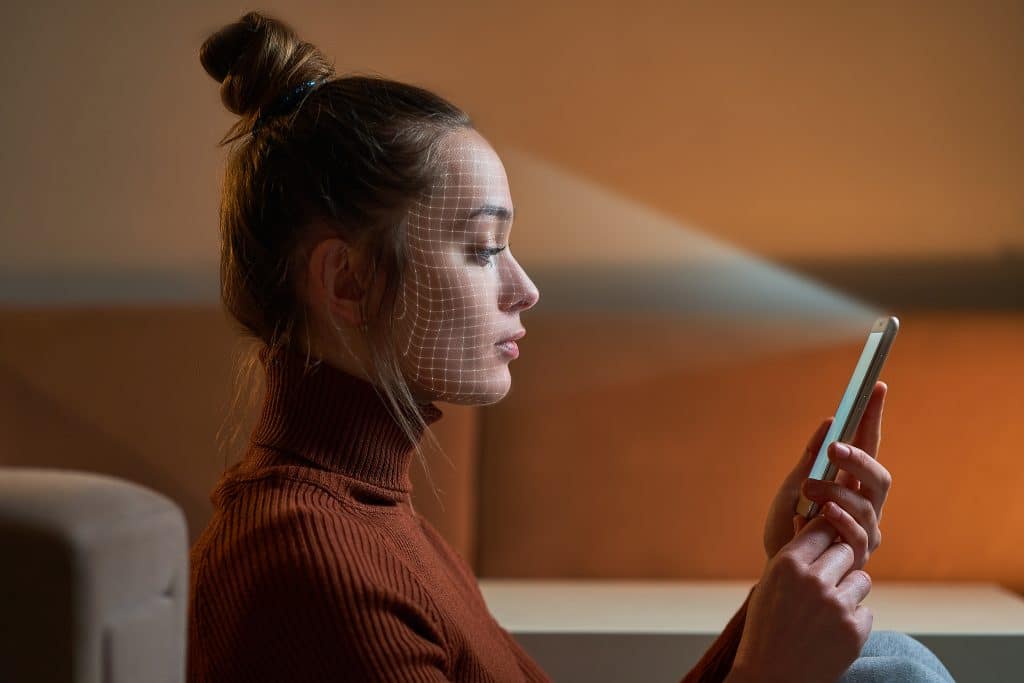 A woman sits indoors, holding a smartphone in front of her. A facial recognition grid is projected on her face, indicating the device is using Face ID to scan her. She has her hair tied up in a bun and is wearing a brown turtleneck sweater.