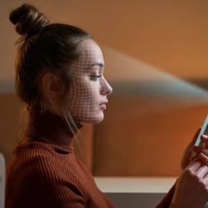 A woman sits indoors, holding a smartphone in front of her. A facial recognition grid is projected on her face, indicating the device is using Face ID to scan her. She has her hair tied up in a bun and is wearing a brown turtleneck sweater.