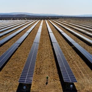 A vast solar farm with rows of solar panels stretching into the distance. In the center, a person in a green shirt walks between the rows. The background features rolling hills under a clear sky, indicating a sunny day and Apple’s commitment to a carbon-neutral supply chain.