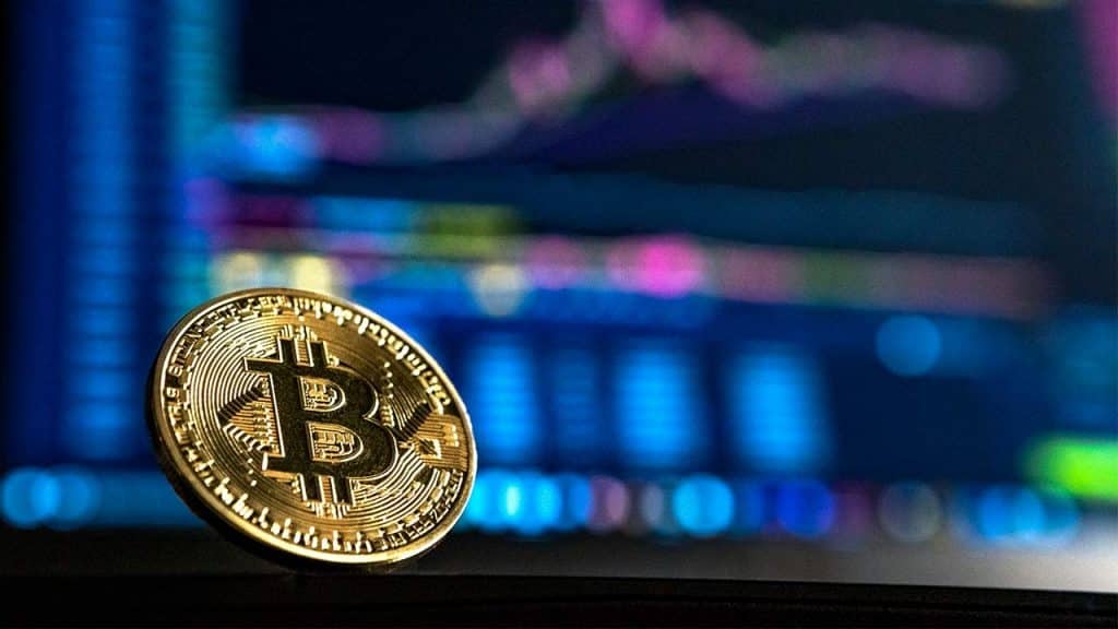 A close-up of a physical Bitcoin coin is in the foreground, with a blurred background of colorful financial graphs and charts displayed on a screen, representing the profitable cryptocurrency market. The Bitcoin symbol is clearly visible on the coin.