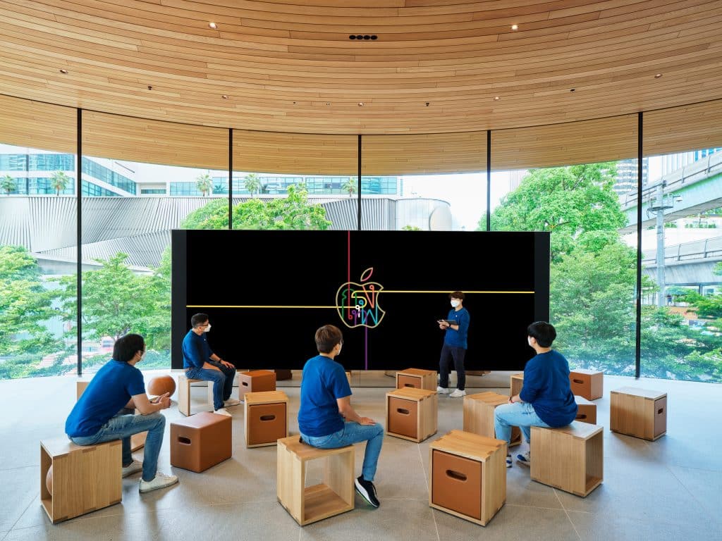 A group of people wearing blue shirts and face masks sit on wooden stools in a modern, circular room with large windows. They face a large screen where an instructor presents. The background shows greenery and a cityscape through the windows of Thailand's new Apple retail store.