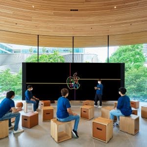 A group of people wearing blue shirts and face masks sit on wooden stools in a modern, circular room with large windows. They face a large screen where an instructor presents. The background shows greenery and a cityscape through the windows of Thailand's new Apple retail store.