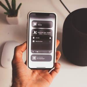 A person holds an iPhone with the home screen open, showing music playback controls on Spotify. Nearby are a HomePod smart speaker, a white wireless mouse, and a small potted plant. The setting appears to be a desk or table, seamlessly blending technology and nature in an iOS 14 environment.