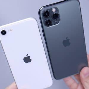 Two hands are holding two iPhones. The iPhone on the left is white, resembling the iPhone SE, with a single camera. The iPhone on the right is black with three cameras. Both have the Apple logo in the center. In the background, a blurry green plant and a white object are visible—a scene familiar to Android users too.