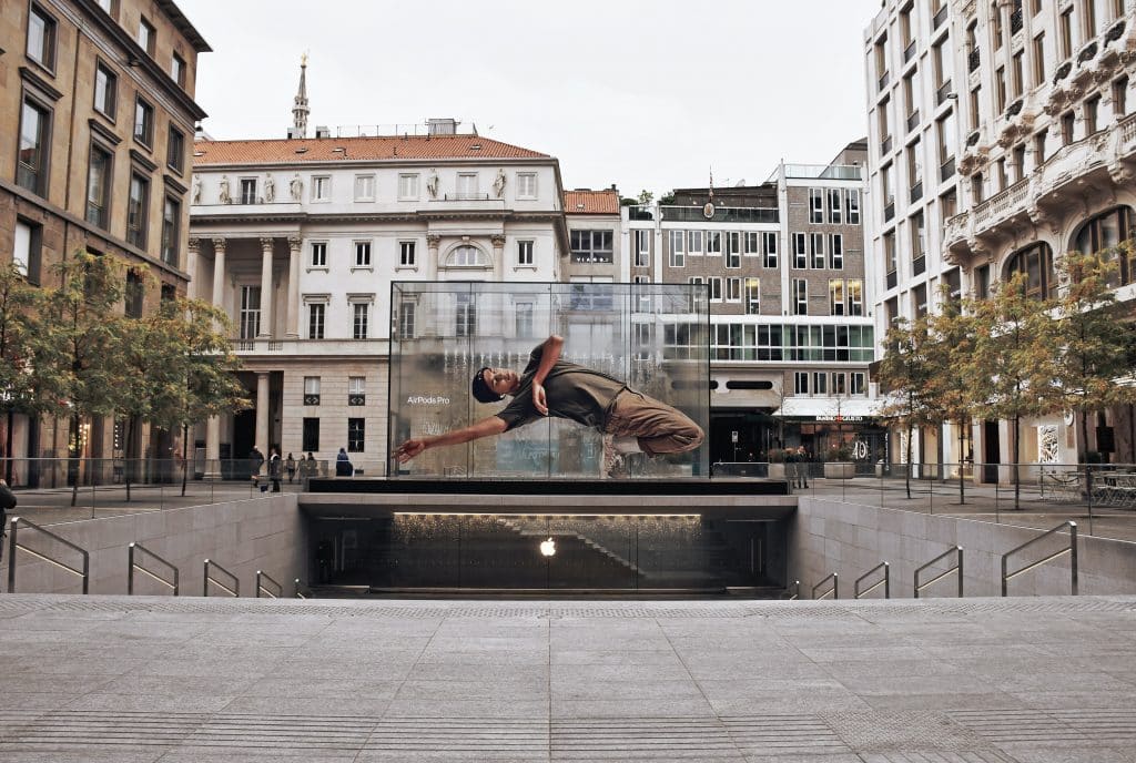 A modern square surrounded by historic buildings features a large, transparent, cube-shaped display with a dynamic image of a person mid-dance. Trees and stairs leading underground enhance the urban setting where visitors can also explore third-party resellers of Apple products.