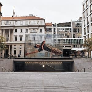A modern square surrounded by historic buildings features a large, transparent, cube-shaped display with a dynamic image of a person mid-dance. Trees and stairs leading underground enhance the urban setting where visitors can also explore third-party resellers of Apple products.