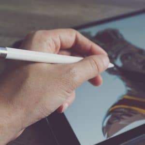 A hand holding an Apple Pencil is drawing a digital illustration of a bearded man on a tablet. The man in the drawing is wearing a yellow and brown outfit, capturing real-world colors perfectly.