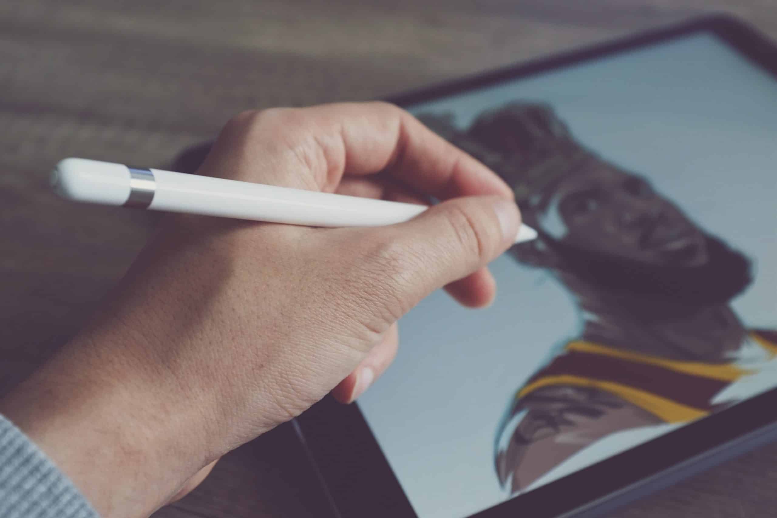 A hand holding an Apple Pencil is drawing a digital illustration of a bearded man on a tablet. The man in the drawing is wearing a yellow and brown outfit, capturing real-world colors perfectly.