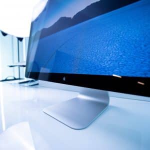 A sleek, modern computer monitor displays a scenic image of a blue landscape with hills in the background. To the left, an Apple laptop with Thunderbolt ports sits on the same reflective white desk. A bright light source illuminates the area from the back.