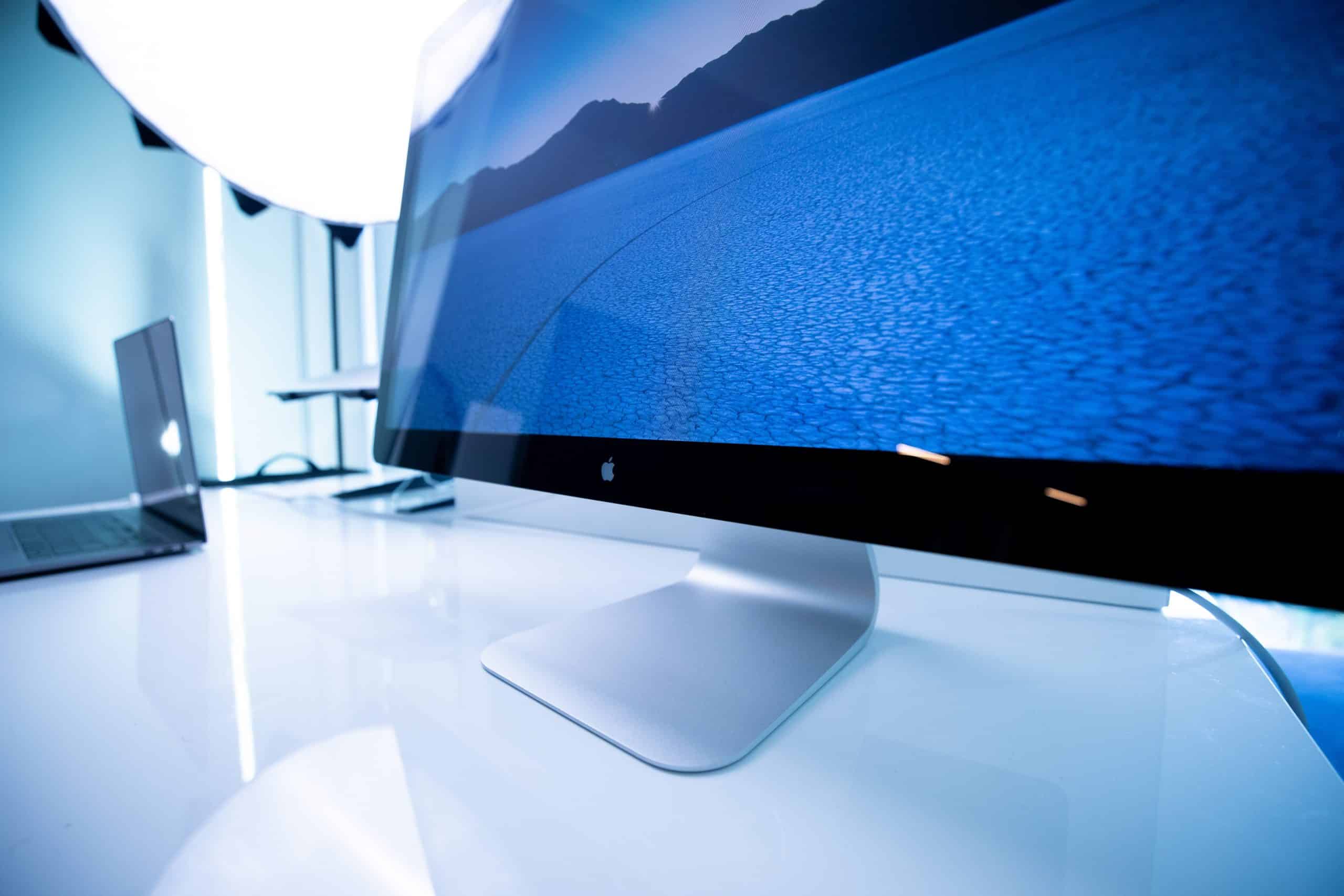 A sleek, modern computer monitor displays a scenic image of a blue landscape with hills in the background. To the left, an Apple laptop with Thunderbolt ports sits on the same reflective white desk. A bright light source illuminates the area from the back.