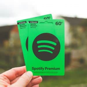 A hand holds two green Spotify Premium gift cards, proudly displaying the Spotify logo. Each card offers six months of Premium service for 60€, providing access to a platform rich with music and exclusive video podcasts. The blurred background hints at an outdoor setting.