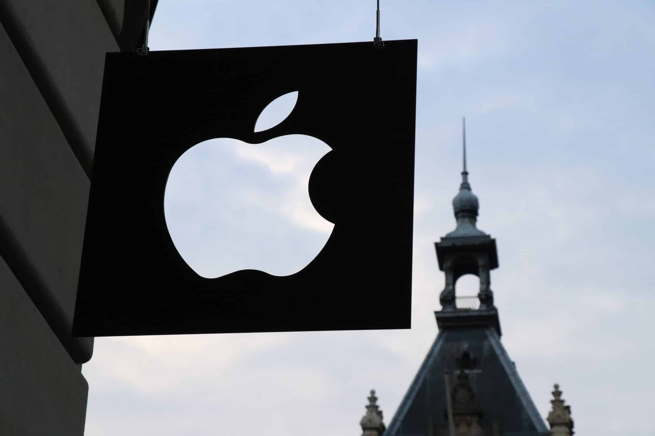 A black sign with a white Apple logo hangs against the backdrop of a historic building with a pointed spire and intricate architectural details. Amid concerns like iPhone battery throttling, the overcast sky adds to the atmosphere.