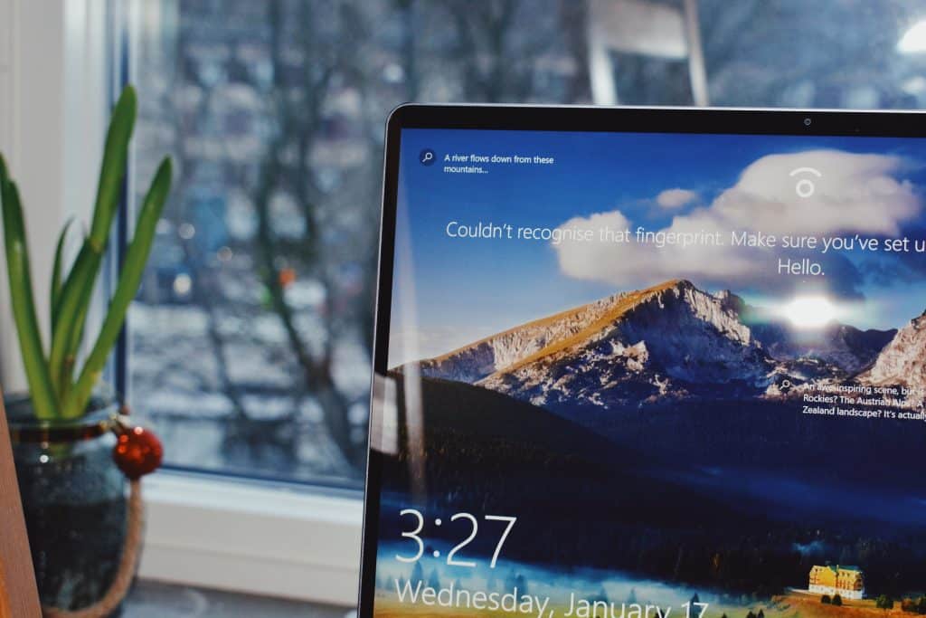 A laptop screen displaying a scenic mountain landscape erroneously indicates an unrecognized fingerprint. The screen shows the time as 3:27 on Wednesday, January 17. In the background, a window reveals a blurred outdoor view with plants on the windowsill, hinting at a new release of Apple software.