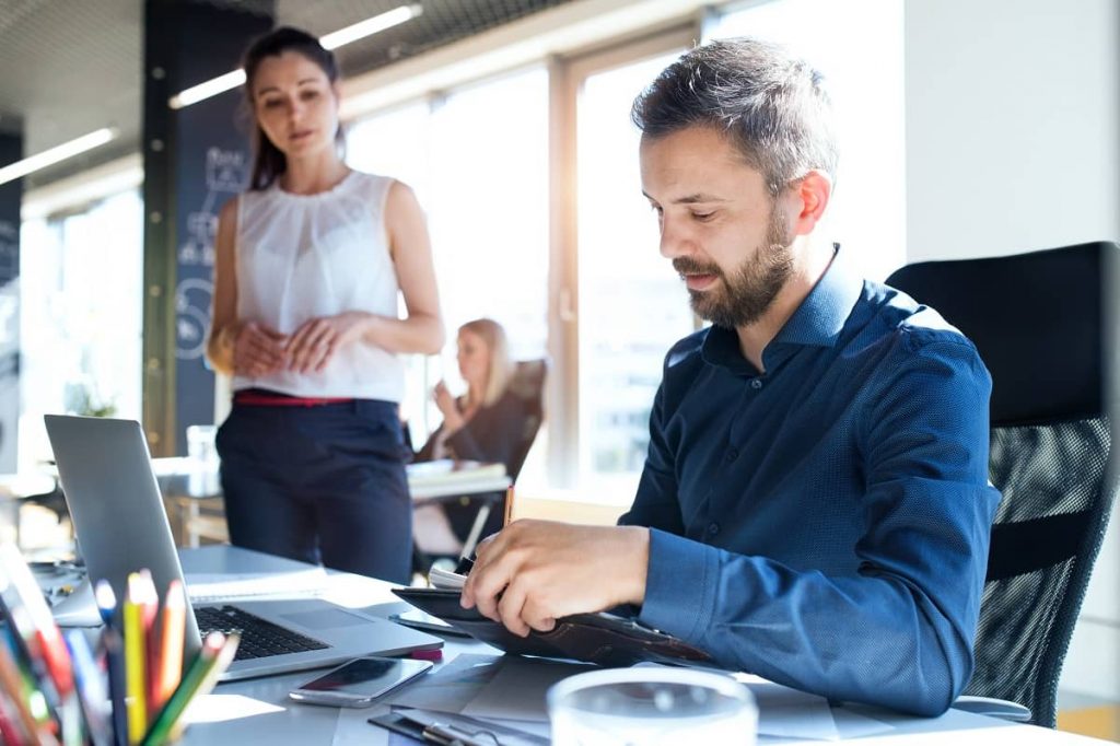 A man in a blue shirt sits at a desk working on a tablet, with a laptop open beside him. A woman in a white sleeveless blouse stands nearby, looking at him. A cup of coffee, colorful pens, and a smartphone are on the desk. Sunlight streams in through the large windows—it's the right time to invest in SEO services.