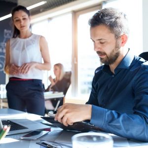 A man in a blue shirt sits at a desk working on a tablet, with a laptop open beside him. A woman in a white sleeveless blouse stands nearby, looking at him. A cup of coffee, colorful pens, and a smartphone are on the desk. Sunlight streams in through the large windows—it's the right time to invest in SEO services.