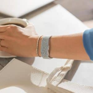 A person wearing a blue shirt and an Apple Watch reaches for a cup of coffee on a white table. Nearby are a white textured bag, a patterned cloth with red hearts, and a plate with pastries.