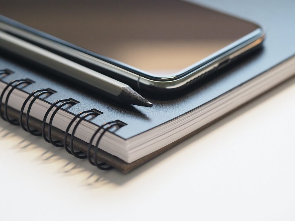 A close-up image of a smartphone, a pencil, and a spiral-bound notebook. The smartphone, showcasing top iOS apps, is placed on top of the closed notebook with the pencil lying next to it. The focus highlights the sleek design of the iOS device and details of the notebook's binding.