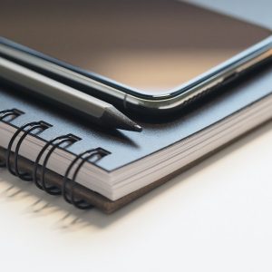 A close-up image of a smartphone, a pencil, and a spiral-bound notebook. The smartphone, showcasing top iOS apps, is placed on top of the closed notebook with the pencil lying next to it. The focus highlights the sleek design of the iOS device and details of the notebook's binding.