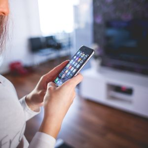 A person is using a smartphone indoors, likely engaged in mobile app testing. The device screen displays various app icons. In the softly lit room with a television and furniture in the background, the focus remains on their hands and the phone as they wear a long-sleeve shirt.