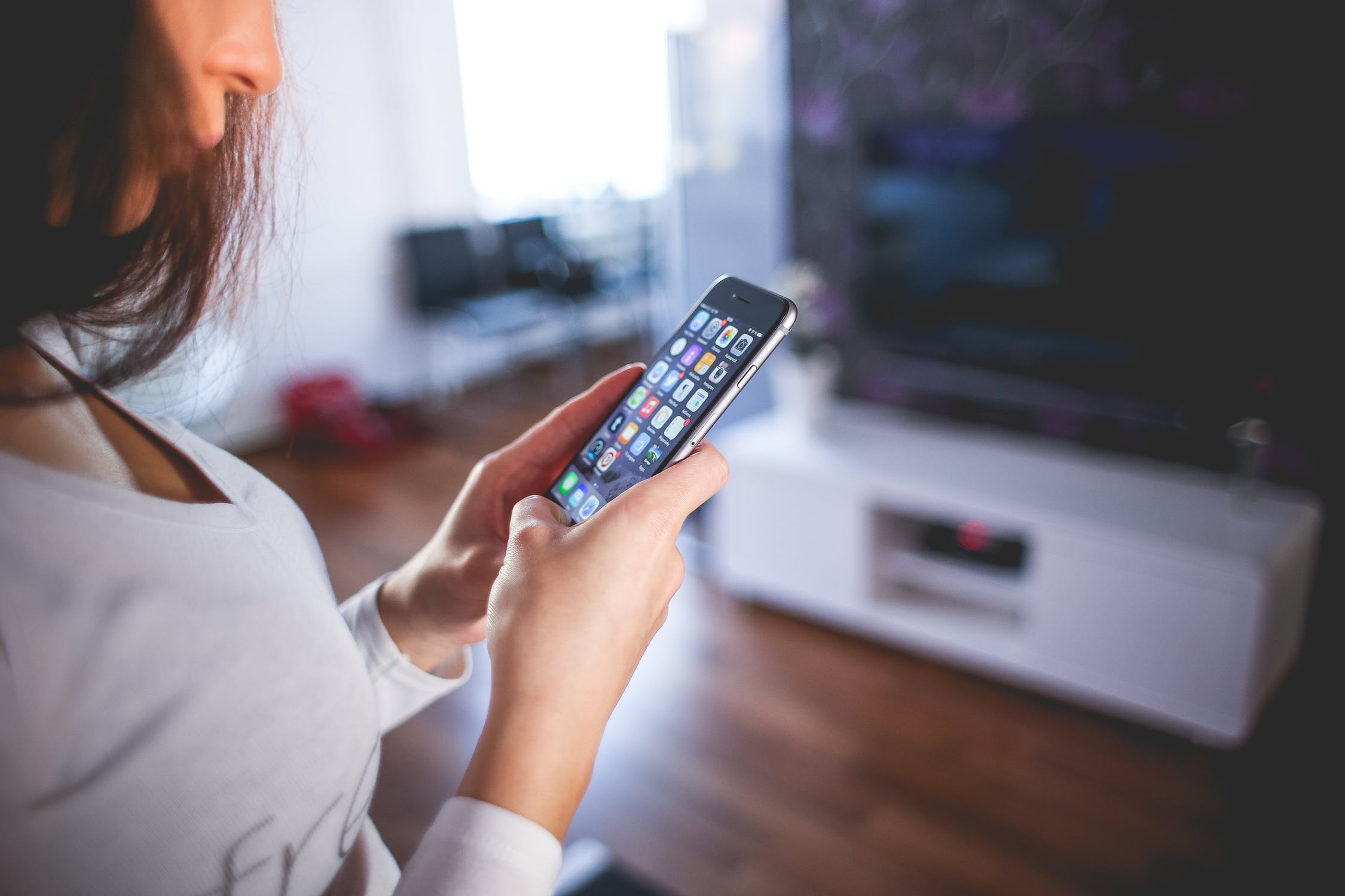 A person is using a smartphone indoors, likely engaged in mobile app testing. The device screen displays various app icons. In the softly lit room with a television and furniture in the background, the focus remains on their hands and the phone as they wear a long-sleeve shirt.