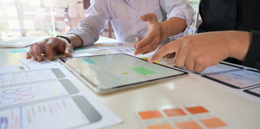 Two students are working collaboratively at a desk covered with papers and digital devices. One is pointing at a tablet displaying a design or prototype through Apple Classroom, while the other is holding a pen and gesturing toward the papers, boosting their learning with various design templates visible on the table.