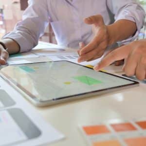 Two students are working collaboratively at a desk covered with papers and digital devices. One is pointing at a tablet displaying a design or prototype through Apple Classroom, while the other is holding a pen and gesturing toward the papers, boosting their learning with various design templates visible on the table.