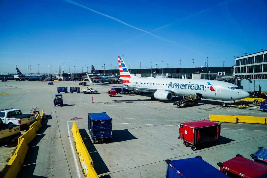 An American Airlines airplane is stationed at an airport gate, surrounded by ground support vehicles and equipment. The sky is clear with faint contrails, and additional planes are visible in the background. Nearby, a traveler watches Apple TV+ as they await their flight.