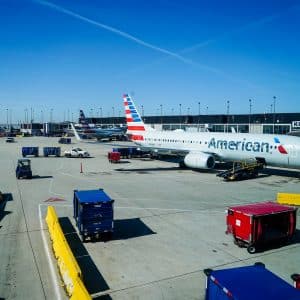 An American Airlines airplane is stationed at an airport gate, surrounded by ground support vehicles and equipment. The sky is clear with faint contrails, and additional planes are visible in the background. Nearby, a traveler watches Apple TV+ as they await their flight.