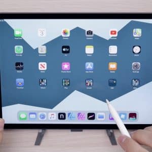 A person is using a stylus with an iPad displaying the home screen, which features various app icons like FaceTime, Calendar, Photos, Camera, Mail, Notes, and more on a blue mountains-patterned wallpaper. This low-cost Apple iPad rests on a wooden surface and includes Touch ID for added security.
