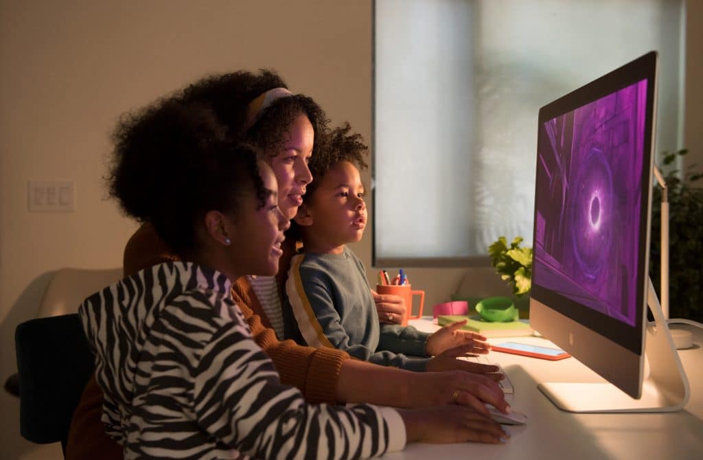 A woman and two children are sitting together at a desk, engaged with a new 27-inch iMac. The screen displays a vibrant, purple spiral pattern. The room is dimly lit, creating a cozy atmosphere while they interact with everything you need to know about the computer.