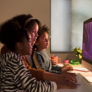 A woman and two children are sitting together at a desk, engaged with a new 27-inch iMac. The screen displays a vibrant, purple spiral pattern. The room is dimly lit, creating a cozy atmosphere while they interact with everything you need to know about the computer.