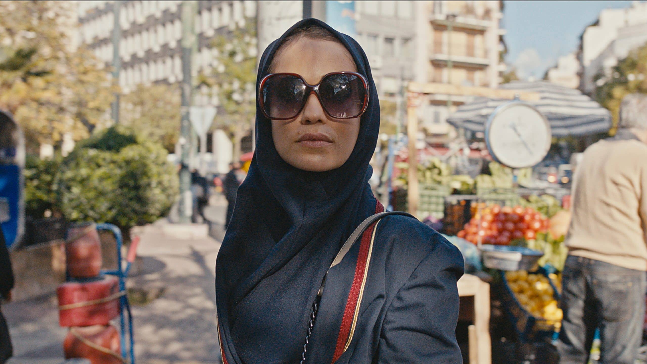 A person wearing oversized sunglasses and a headscarf walks through an outdoor market, reminiscent of a scene from an Israeli series on Apple TV+. In the background, there are various market stalls with fruits and vegetables, a scale, greenery, and people. The day appears to be sunny.