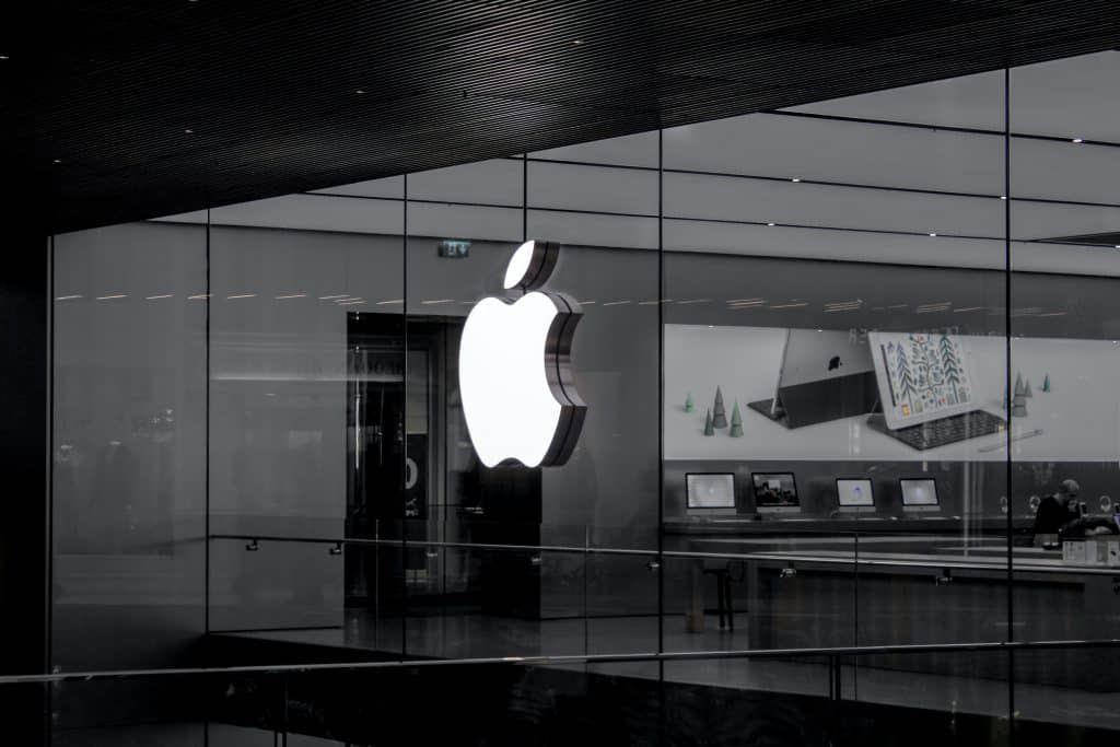 A large, illuminated Apple logo is displayed on the glass wall of a modern retail store in India. Inside, various Apple products, such as laptops and accessories, are showcased on tables. The interior is sleek and minimalist, with bright lighting and a clean design reminiscent of their online retail store.