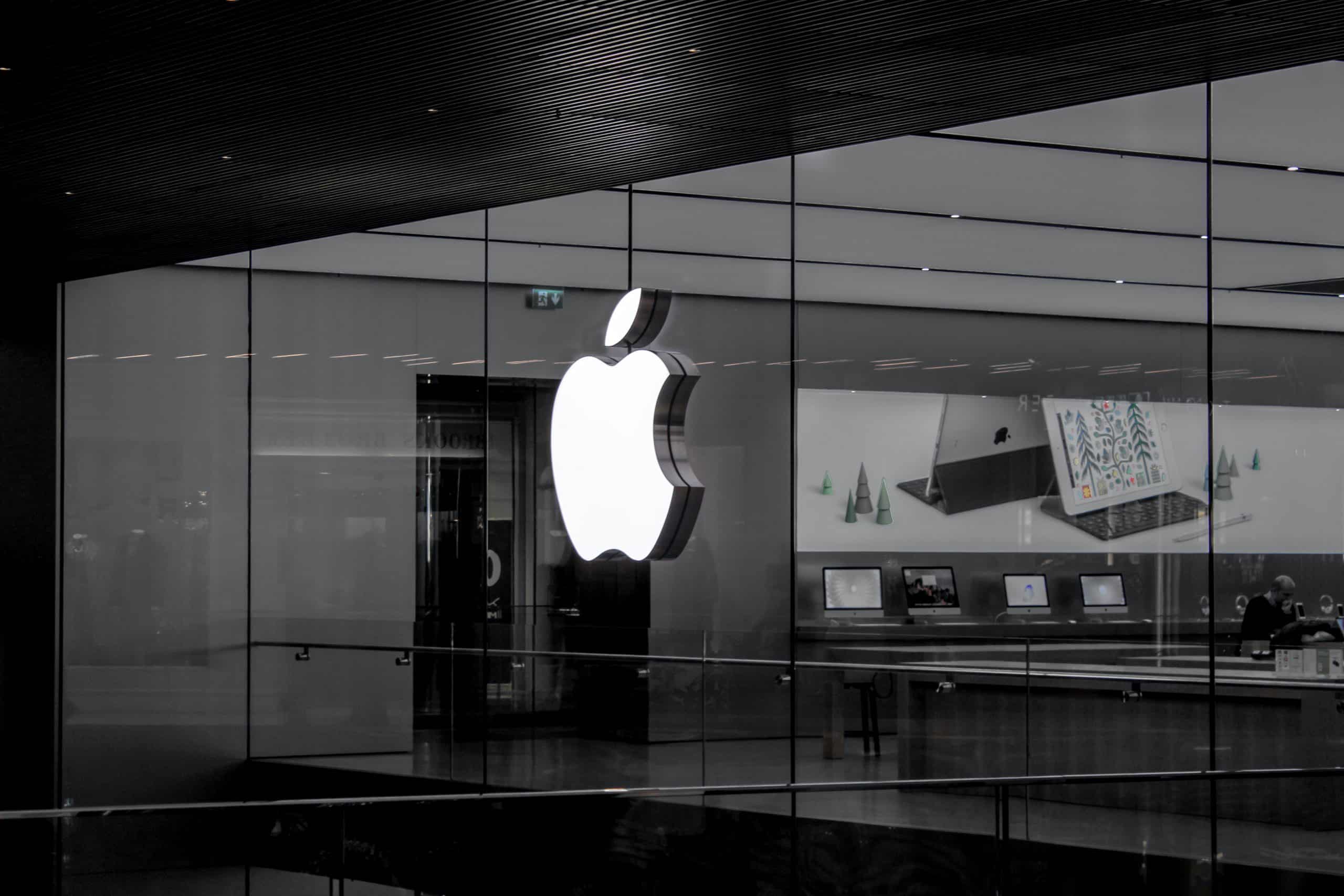 A large, illuminated Apple logo is displayed on the glass wall of a modern retail store in India. Inside, various Apple products, such as laptops and accessories, are showcased on tables. The interior is sleek and minimalist, with bright lighting and a clean design reminiscent of their online retail store.