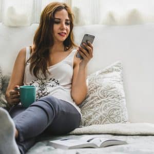 A woman with long brown hair is sitting on a cushioned surface, leaning against pillows. She is holding a green mug in one hand and looking at her smartphone in the other hand, likely exploring dating apps. An open book lies on the cushion near her feet. Daylight illuminates the scene.