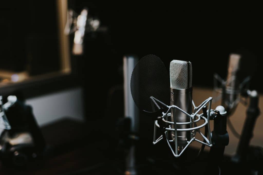 A close-up of a professional condenser microphone with a pop filter in a dimly lit recording studio, ideal for capturing the high-quality audio required for a premium podcast. Another microphone and some studio equipment are blurred in the background.