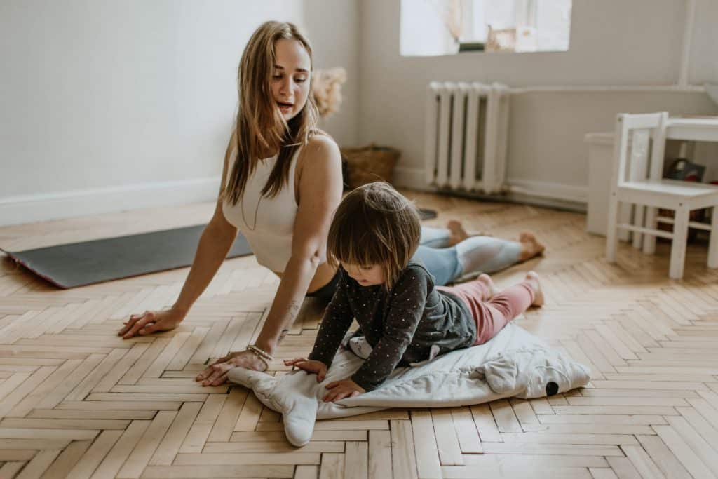 A woman and a child are practicing yoga on a wooden floor in a bright room. The woman stretches while sitting on one knee, looking at the child, who lies on a blanket, also stretching. A yoga mat and a small table and chairs are visible in the background. They’re enjoying their new Apple Fit fitness subscription launched in October.