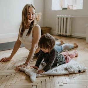 A woman and a child are practicing yoga on a wooden floor in a bright room. The woman stretches while sitting on one knee, looking at the child, who lies on a blanket, also stretching. A yoga mat and a small table and chairs are visible in the background. They’re enjoying their new Apple Fit fitness subscription launched in October.