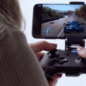 A person is holding a black gaming controller attached to an iPhone displaying a racing game. The game shows a car racing on a scenic road with trees and mountains in the background. The person's hands are visible, immersed in controlling the action through Microsoft’s xCloud.