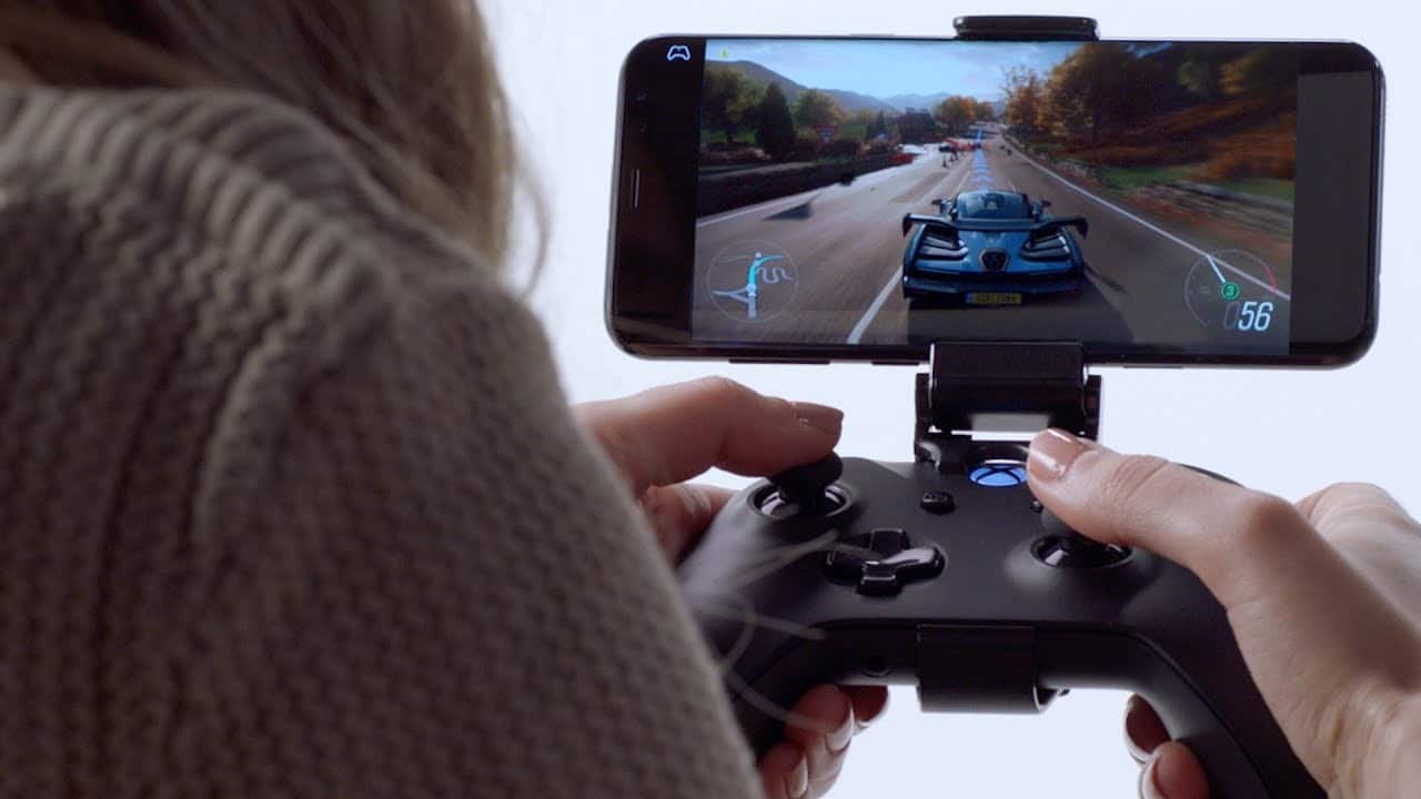 A person is holding a black gaming controller attached to an iPhone displaying a racing game. The game shows a car racing on a scenic road with trees and mountains in the background. The person's hands are visible, immersed in controlling the action through Microsoft’s xCloud.