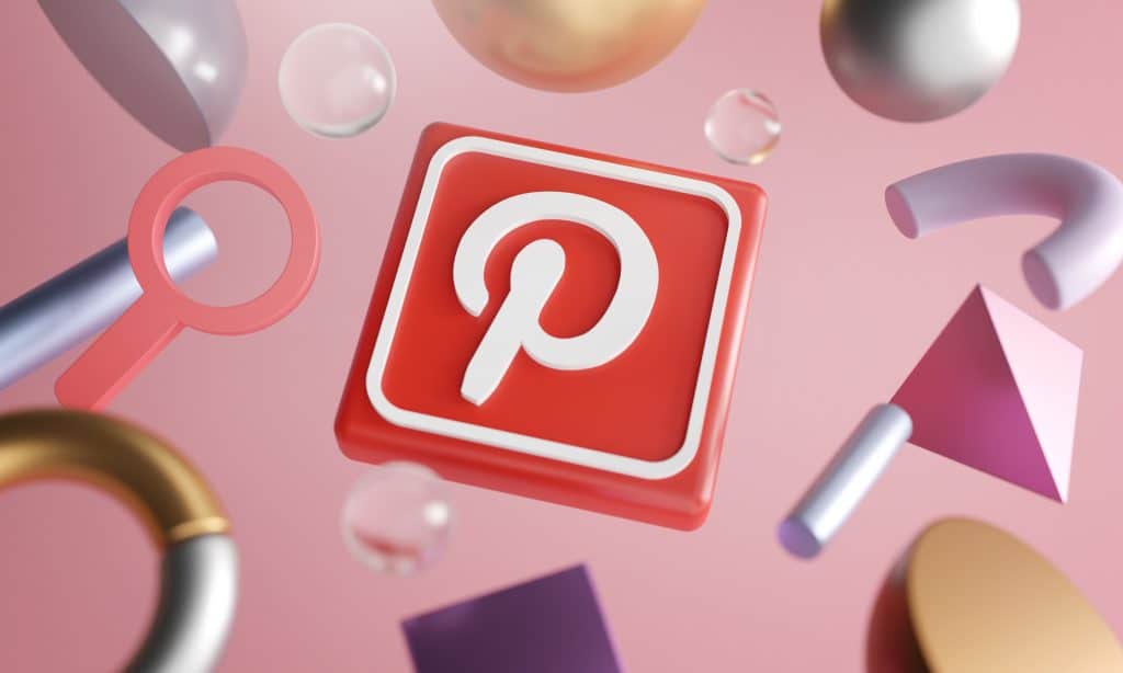 A red 3D Pinterest logo surrounded by various floating geometric shapes, including spheres, cubes, and curves, all set against a pink gradient background. The colorful shapes enhance the creative and dynamic feel of an image reminiscent of iOS 14’s vibrant App Store design.