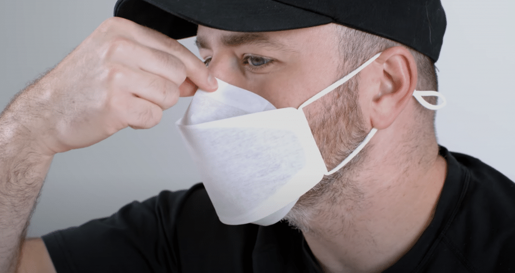 A man wearing a black cap and a black shirt is adjusting his white, Apple-branded face mask. He is using his right hand to press the mask against the bridge of his nose. The mask covers his nose and mouth securely.