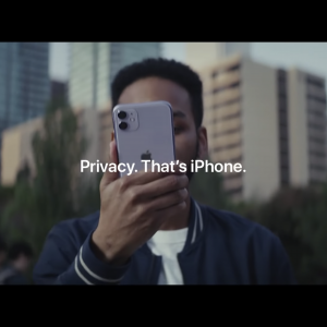 A person standing outdoors in a city environment holds up a silver iPhone, covering half of their face. The background shows blurred buildings and trees. Text in the center of the image reads, "Privacy. That's iPhone," showcasing Apple’s focus on being a privacy-focused smartphone brand.